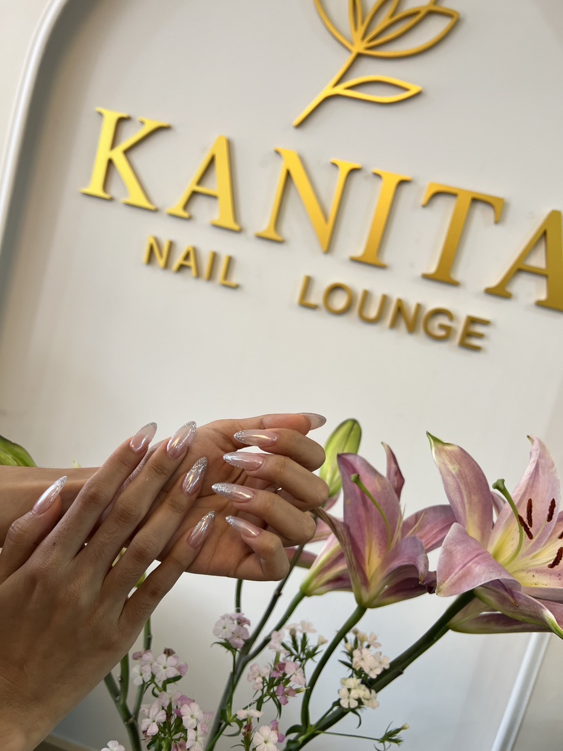 Soft pink chrome manicure photographed with flowers and the Kanita Nail Lounge logo